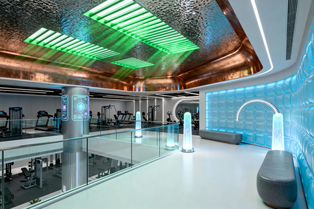 A spacious, two-level futuristic home gym features a white mezzanine with clear glass railings overlooking cardio and weight machines below. Overhead, a hammered copper ceiling wraps around green LED slots that wash light across the space. A wall of blue backlit glass blocks frames a seating niche with two gray benches and glowing acrylic totems linked by a segmented arc light. Linear LEDs, smooth white floors, and matte black equipment create a crisp, high-tech atmosphere.