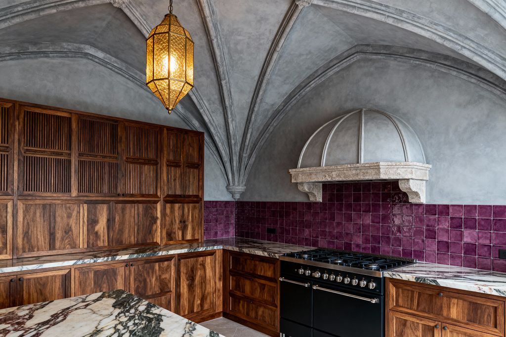 Kitchen with ribbed, pointed vaults in gray plaster and a domed carved-stone range hood above a black range. Walnut cabinets with slatted uppers and veined marble countertops wrap an L-shaped corner against a plum square-tile backsplash, lit by a faceted brass lantern with textured glass, while the floors are pale stone and the overall palette blends warm wood, deep purple, and smoky gray.