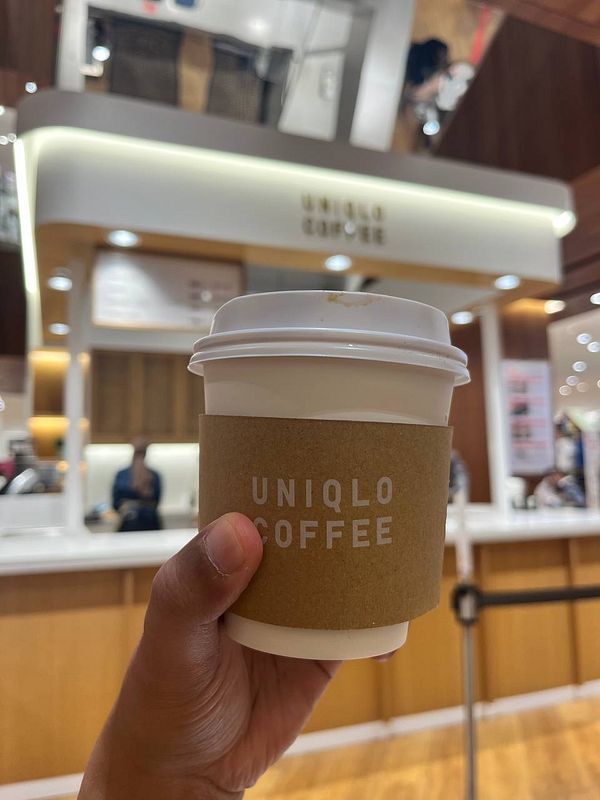 A hand holds a cup of Uniqlo coffee in front of the Uniqlo Coffee counter.