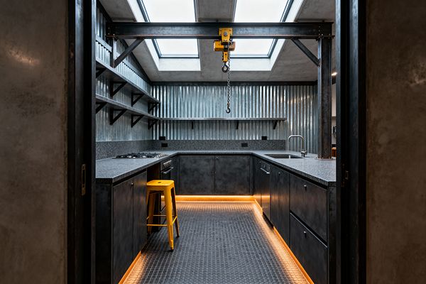 U-shaped industrial kitchen clad in corrugated galvanized steel with blackened steel cabinets and speckled gray countertops. Exposed steel I-beams frame two skylights and suspend a yellow hoist with chain and hook; open shelves with black brackets line the left wall; a stainless sink sits to the right; a gas cooktop and oven anchor the left; coin-pattern rubber flooring and warm toe-kick LEDs rim the base, with a yellow metal stool as an accent.