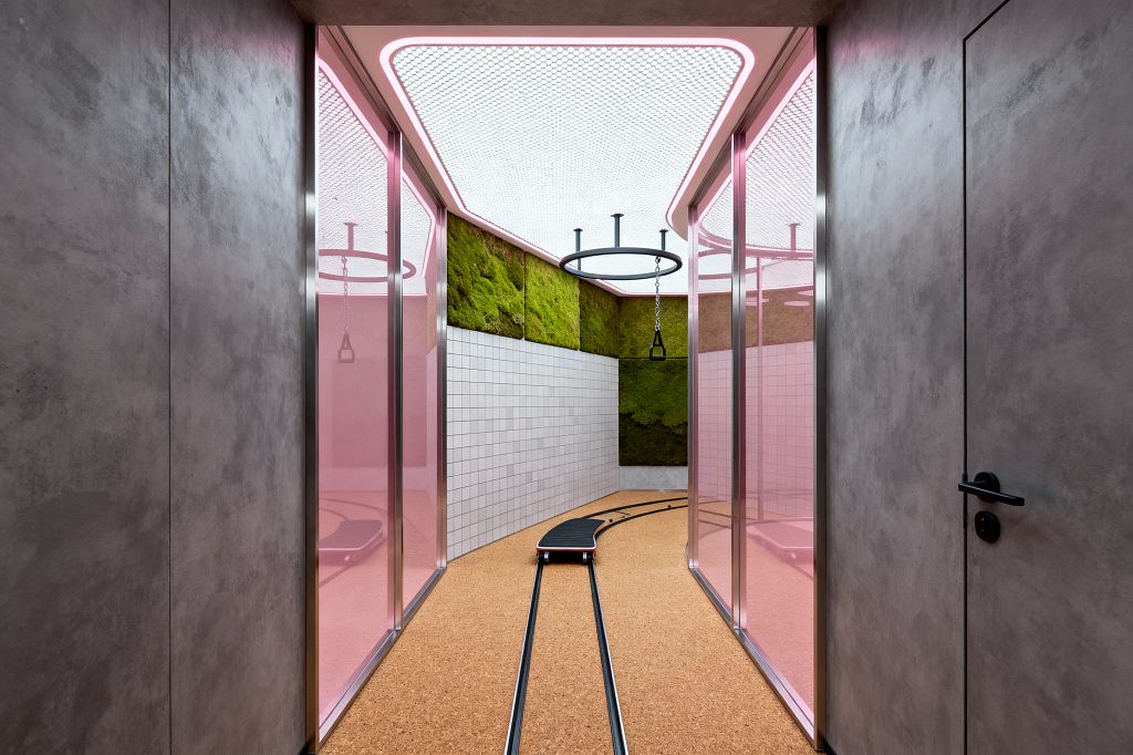 A narrow corridor with gray microcement walls opens to a compact home gym enclosed by pink-tinted glass panels in stainless frames. Inside, warm cork flooring integrates twin metal rails with a small wheeled board, while walls combine white square ceramic tile and preserved moss panels; above, a white perforated mesh ceiling edged in neon-pink LED light supports a circular steel rig with suspended triangle handles, creating a sleek, sci‑fi ambience.