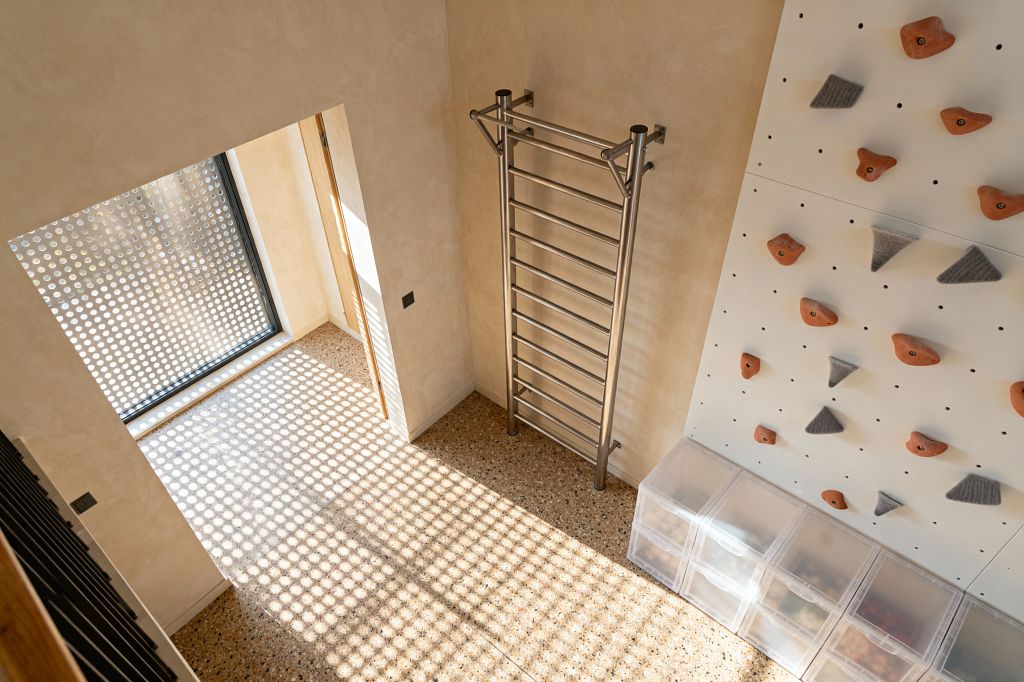 Overhead view of a compact modern home gym with a white climbing wall fitted with terracotta-orange and gray holds, a brushed stainless steel stall bar mounted on a beige plaster wall, and speckled terrazzo flooring. Daylight pours through a large window shaded by a perforated metal screen, casting a grid of circular light across the floor; clear plastic storage bins line the base of the wall, while a black switch and a slim wood-trimmed doorway complete the minimal space.