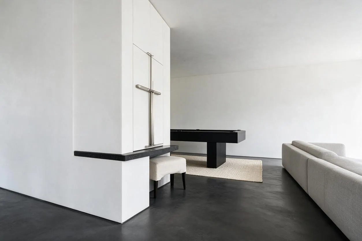 A minimalist game room with white walls, a dark polished floor, and a black pool table centered on a light woven rug. A cream upholstered sofa sits along the right side, while a small upholstered stool and a built-in white column with a metal cross-like detail add sculptural simplicity.