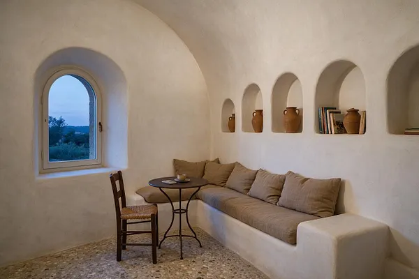 A compact Mediterranean-style studio apartment with white stucco walls, a vaulted ceiling, and a series of built-in wall niches holding clay vases and a few books. A long built-in bench with beige cushions runs along the wall beside a small round café table and a woven-seat wooden chair, while an arched window brings in soft blue dusk light.