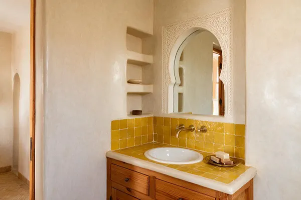 A Moroccan-style powder room with plaster walls, a carved arched mirror, and a compact vanity topped with glossy mustard-yellow tile. The white oval sink sits beneath brass wall-mounted faucets, with a wood cabinet below and built-in wall niches beside the mirror.