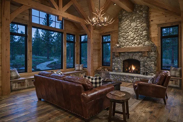 A rustic backyard lounge with a vaulted wood ceiling, exposed beams, and a large stone fireplace centered on the far wall. Leather seating, log-style benches, and floor-to-ceiling windows overlook a wooded landscape and winding stone path.