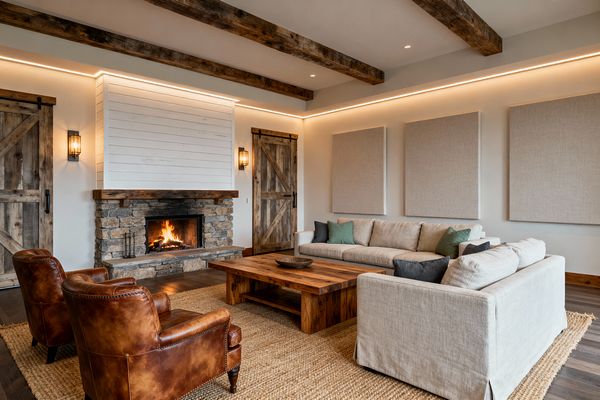A farmhouse-inspired media room features a stacked-stone fireplace with a thick reclaimed-wood mantel and white shiplap above. A beige linen sectional surrounds a chunky reclaimed-wood coffee table, paired with two cognac leather club chairs on a jute rug over dark wood floors. Three large fabric wall panels sit opposite the hearth, while rustic sliding barn doors flank it. Exposed timber beams, warm LED cove lighting, recessed spots, and metal sconces create an inviting glow.