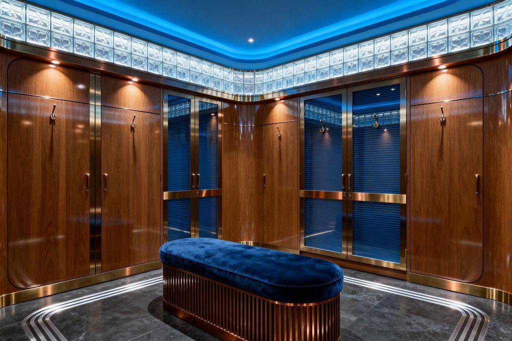 A luxurious Art Deco mudroom lined with walnut cabinets featuring rounded corners and satin brass trim. Reeded blue glass doors in brass frames, brass coat hooks, and integrated spotlights add shine. A band of translucent glass blocks and blue cove lighting wraps the ceiling. Centered is an oval bench upholstered in deep navy velvet on a slatted metallic base. The floor is dark stone with curved metal inlays, reinforcing the streamlined, glamorous aesthetic.