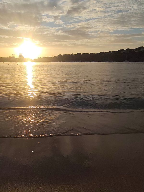 Sunset over a calm beach with gentle waves and a silhouette of boats and land in the background.