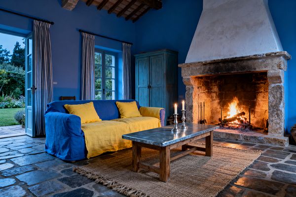 A French Country living room features cobalt-blue walls and matching French doors with striped linen curtains, centered on a large limestone fireplace with a roaring fire. A blue linen slipcovered sofa layered with sunflower-yellow pillows and throw faces a zinc-topped wood coffee table over a chunky jute rug on flagstone floors; a distressed turquoise armoire and exposed timber beams with terracotta ceiling tiles frame the space, while silver candlesticks and iron fire tools add metallic accents, and a garden lies just outside.
