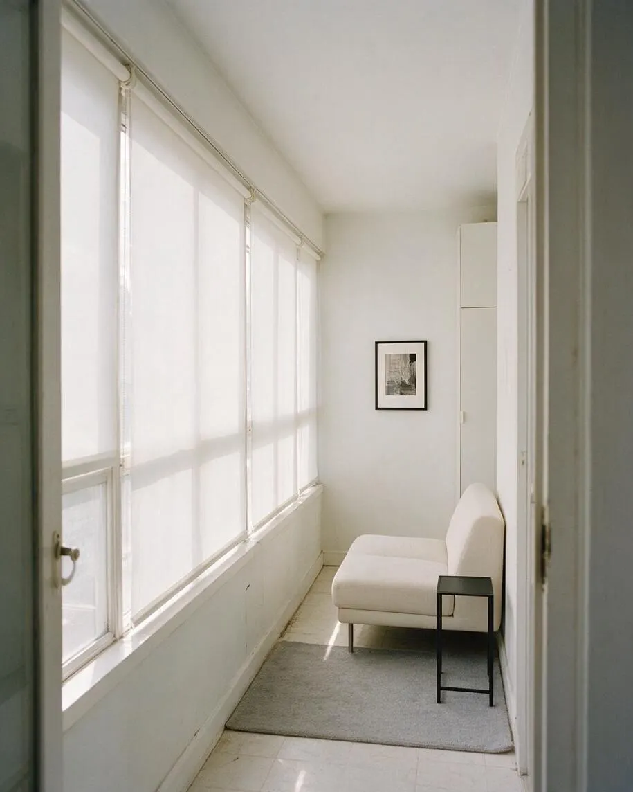 A narrow minimalist sunroom with large floor-to-ceiling windows on the left covered by sheer white shades, filling the space with diffused light. The room features a small white chaise lounge, a black side table, a light gray rug, and a framed black-and-white artwork on the back wall, all set against crisp white walls.