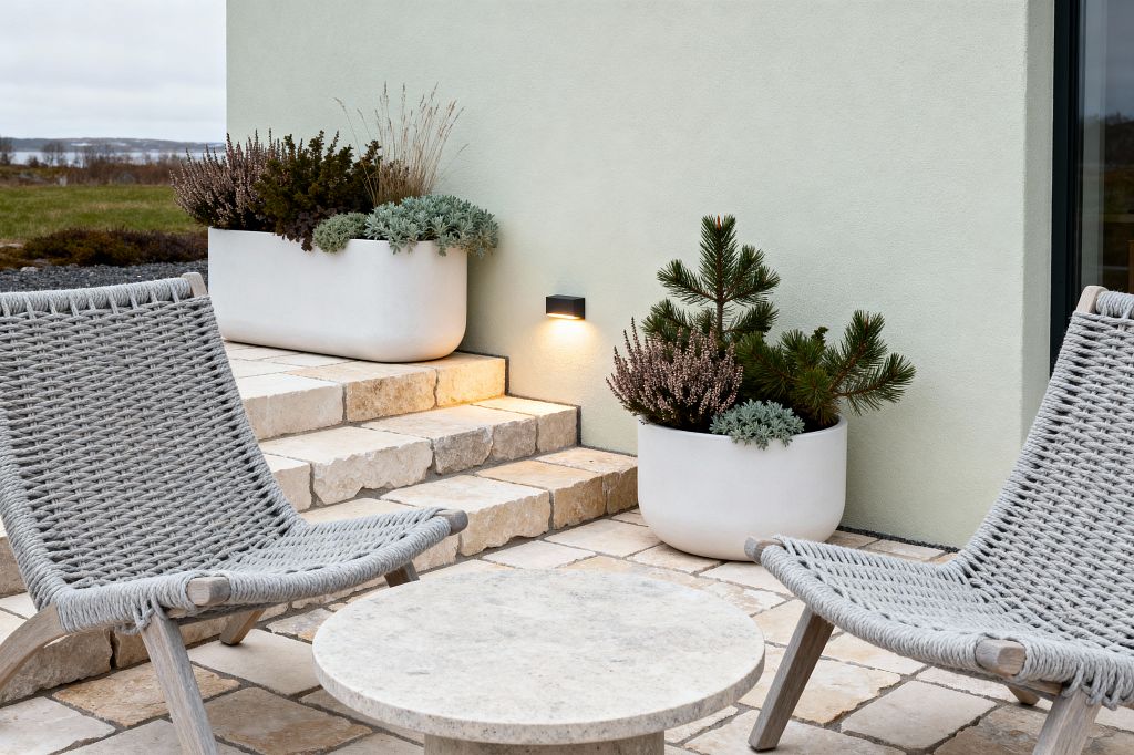 A Scandinavian patio featuring two low gray rope-woven lounge chairs with light wood frames, a round light-stone coffee table, and tumbled limestone pavers and steps leading to a pale sage plaster wall. Matte white planters with evergreen pine, heather, grasses, and succulents flank a warm LED wall sconce, with open landscape beyond.