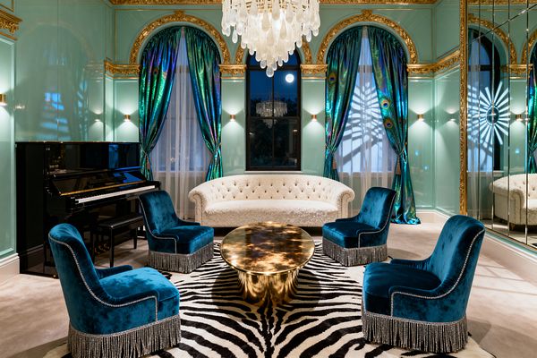 Opulent Hollywood Regency living room with mint lacquered walls, ornate gilded arches, mirrored panels, and a cascading white glass chandelier. A curved tufted bouclé sofa faces four peacock-blue velvet chairs with silver fringe around a polished brass round coffee table on a zebra rug, framed by iridescent peacock-pattern drapes, soft wall sconces, and a glossy black upright piano.