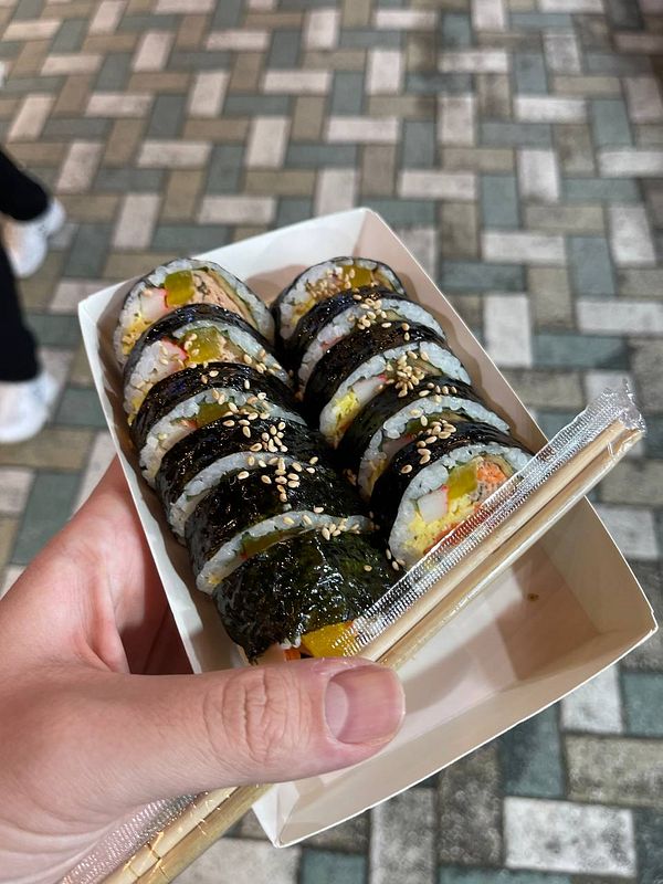 A hand holds a container of sushi rolls against a patterned pavement.