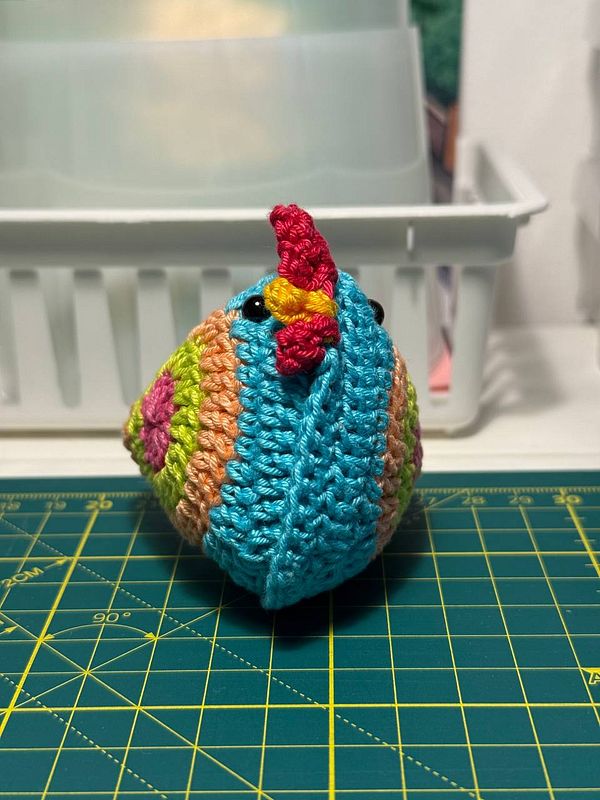 A colorful crocheted critter resembling a bird is displayed on a cutting mat.