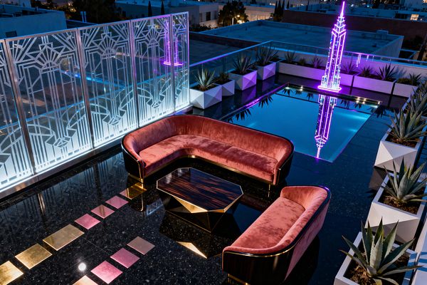 Nighttime view of a rooftop patio in Art Deco style. Two curved sofas in dusty pink velvet with brass trim face a faceted dark-wood and brass coffee table on a glossy black terrazzo floor with pink and gold inlays. A reflecting pool mirrors a neon magenta spire sculpture. Backlit frosted glass panels show geometric patterns, and rows of agave plants sit in angular white planters behind a glass railing overlooking the city.