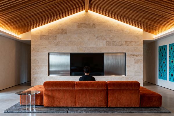 A Mid-Century Modern media room features a vaulted ceiling of narrow wood slats with concealed cove lighting, focusing on a travertine-clad wall with a recessed television framed by brushed metal panels. A burnt-orange corduroy sectional sits on a dark rug over polished concrete alongside a clear acrylic side table, with teal graphic art panels to the right and open hallways flanking the central wall.