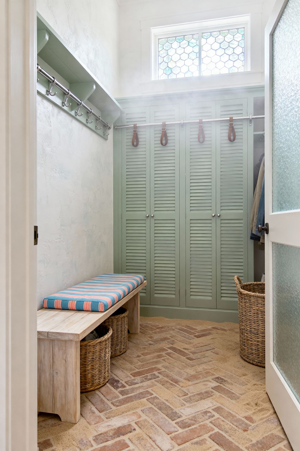 A compact coastal mudroom features seafoam green louvered lockers on a stainless rail with leather pulls and small round knobs. A light-washed wood bench with a turquoise and coral striped cushion sits opposite a shelf and row of metal hooks, with woven baskets below; herringbone brick flooring and sandy grout lead to a frosted glass door, while light filters through a hex-pattern clerestory window for an airy glow.