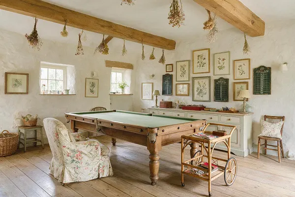 A bright cottage-style game room with a wooden billiards table at the center, floral-upholstered chairs, exposed ceiling beams, and dried flowers hanging overhead. The white plaster walls display botanical prints, while pale wood floors and antique-style furnishings create a warm, rustic atmosphere.