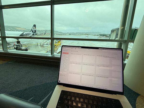 A laptop displaying a task management interface is positioned in an airport lounge with a view of an airplane and runway.