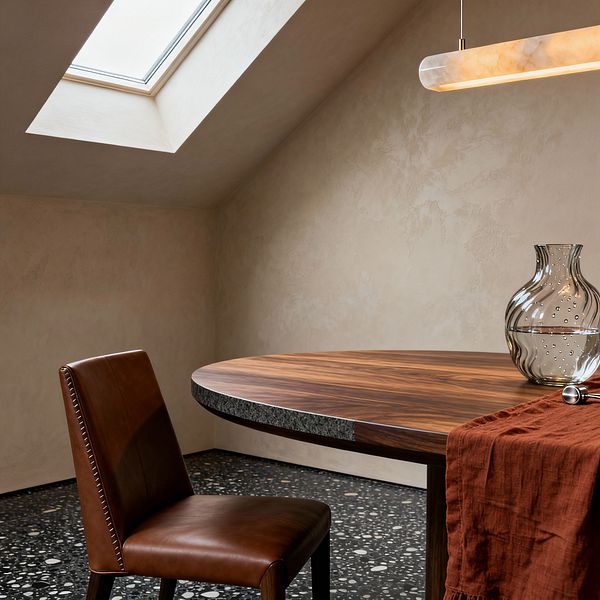 Contemporary dining nook beneath a sloped skylight with lime-washed beige plaster walls and a black terrazzo floor. A round walnut table includes a segment of rough dark stone at the edge. A cognac leather chair with stitched seams sits next to the table. Overhead, a slim alabaster pendant with brass accents glows softly. On the table, a rippled clear glass carafe and a rust linen cloth add texture.