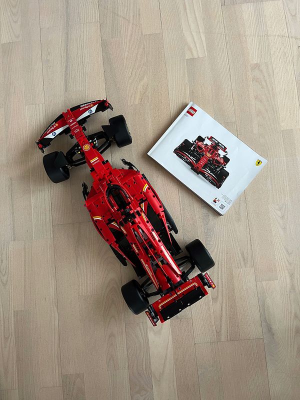 A completed Lego model of a red Formula 1 car is displayed alongside its instruction booklet on a wooden floor.