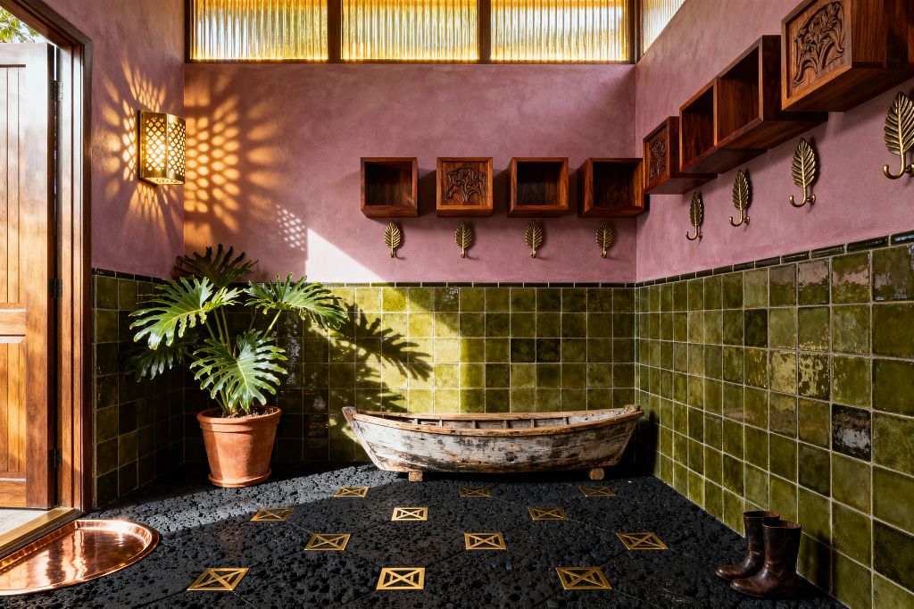 Sunlit tropical mudroom with dusty-pink plaster above olive green square tiles. Textured black lava-stone floor with brass inlays; rustic wooden canoe used as bench; row of brass leaf hooks under carved teak cubbies. Large philodendron in terracotta pot near wooden door; woven rattan sconce casts starburst light; high amber clerestory windows glow.
