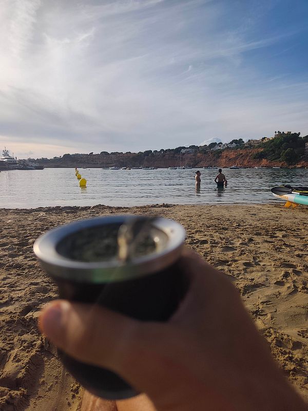 View from a sandy beach looking out over calm water with people swimming and boats anchored near a hilly coastline under a partly cloudy sky, with a hand holding a metal cup in the foreground.
