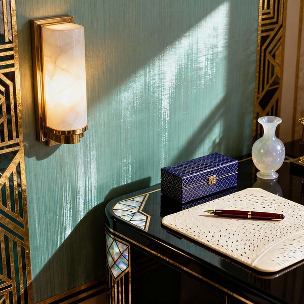 Close-up of a vintage glam home office vignette: a brass wall sconce with an alabaster shade casts warm light on a seafoam green, subtly shimmering wall. A glossy black lacquer desk edged with brass and mother-of-pearl inlay holds a textured cream desk blotter with a burgundy pen, a cobalt blue patterned box with a clasp, and a small opaline glass vase. Sunlight streaks across the surface, highlighting gold geometric trim and reflective finishes.