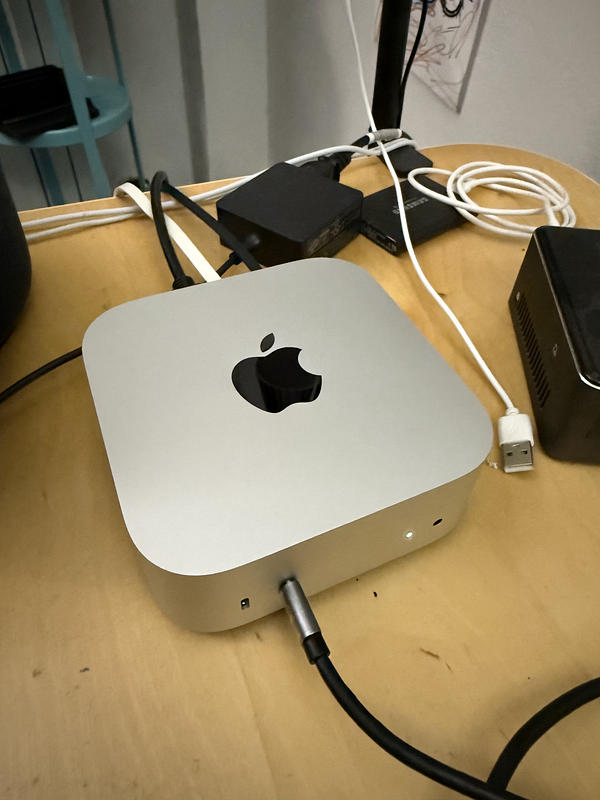 A Mac Mini is prominently displayed on a wooden desk surrounded by various cables and devices.