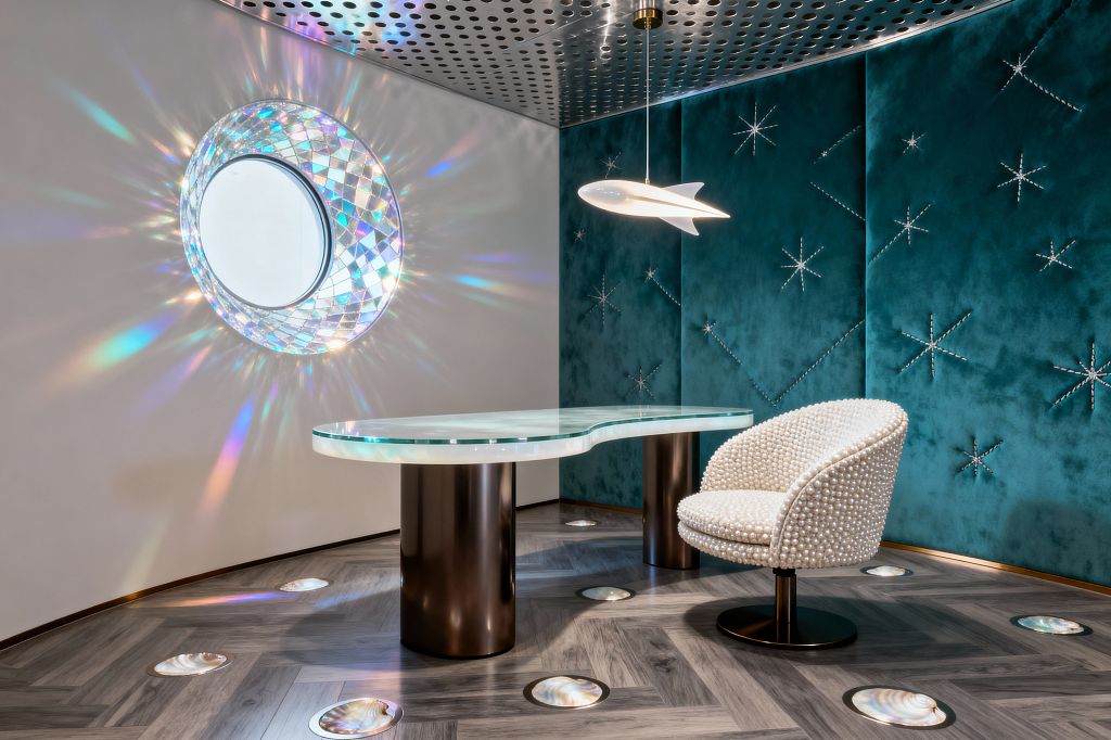 Curved home office with a teal velvet feature wall stitched with starburst constellations, a sculptural kidney-shaped glass desk on bronze cylinder legs, and a rounded chair upholstered in pearl-like beads. A circular prismatic window casts rainbow rays across a warm white wall and gray herringbone floor dotted with iridescent round insets. Overhead, a perforated metal ceiling suspends a white rocket-shaped pendant, creating a luminous, space-inspired atmosphere.