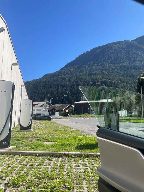 A Tesla vehicle is parked near charging stations with a mountainous backdrop under a clear blue sky.