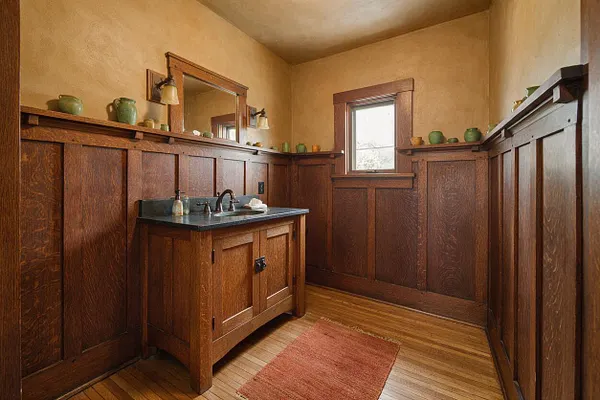 A Craftsman-style powder room with dark stained wood wainscoting and trim, beige textured walls, and a small wood vanity with a dark stone countertop. A mirror and wall sconces sit above the sink, a narrow window brings in daylight, and a muted red rug rests on the wood floor.