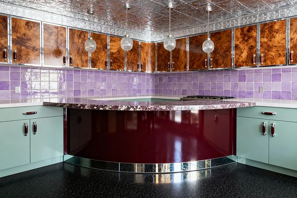 A vintage glam kitchen centers on a curved, high-gloss burgundy peninsula beneath three ribbed glass globe pendants. Lilac ceramic tile lines the backsplash, mint green base cabinets with chrome and maroon pulls sit below glossy burl wood uppers edged in chrome, a pressed-tin silver ceiling and mirrored toe-kick add sparkle, while a purple-flecked stone countertop, gas cooktop, and black speckled floor complete the retro-luxe space.
