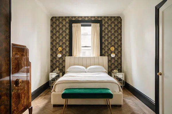 A narrow master bedroom with a cream upholstered bed centered against a dramatic black-and-gold Art Deco accent wall. The room has white walls, a herringbone wood floor, mirrored bedside tables, brass wall sconces, cream curtains, and a green velvet bench at the foot of the bed.