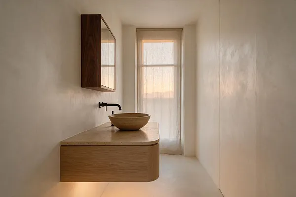 A narrow Japandi-style bathroom with smooth white walls, a floating light wood vanity, and a round stone vessel sink. A dark wall-mounted faucet and matching wood mirrored cabinet add contrast, while a sheer curtain filters warm natural light from the window.