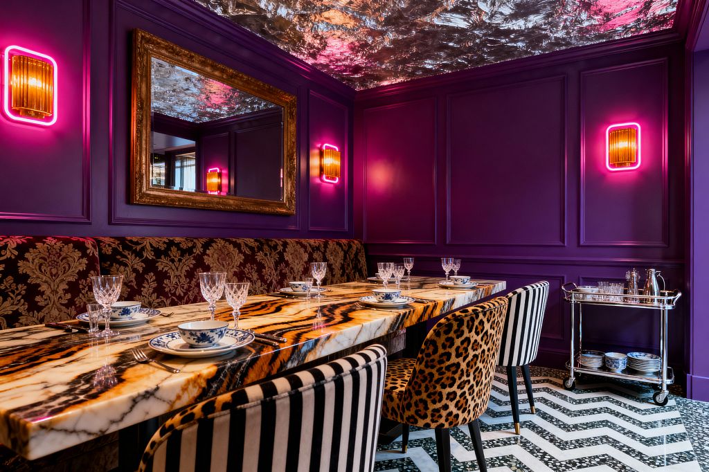 A maximalist dining room features deep purple paneled walls, a reflective silver-leaf ceiling, and neon-framed amber sconces flanking a gilded mirror. A long polished onyx table with bold honey, white, and black veining is paired with a damask banquette, leopard-print and black-and-white striped chairs, blue-and-white porcelain place settings, crystal stemware, a black-and-white chevron tile floor, and a chrome bar cart.