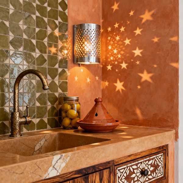 A close-up of a Moroccan-style kitchen corner shows olive green geometric zellige tiles meeting a terracotta tadelakt wall. A perforated metal wall sconce projects star-shaped light patterns over a honed limestone counter with an integrated stone sink, an aged brass gooseneck faucet, a wood drawer with bone-style inlay, and accessories including a clay tagine and a glass jar of preserved lemons.