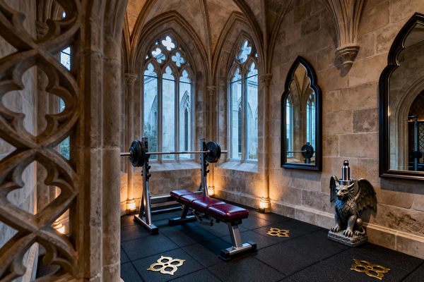 A compact home gym sits inside a Gothic stone room with ribbed vaults, pointed arches, and tall windows filled with quatrefoil tracery. A steel bench press with burgundy leather pads rests on black rubber tiles detailed with gold quatrefoils. Warm uplights wash the columns, two pointed-arch mirrors line one wall, and a carved gargoyle pedestal holds a water bottle, while views of an exterior stone facade appear through the windows.