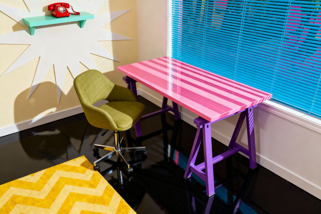 A Pop Art home office features a glossy hot pink desk on purple trestle legs beside a large window with bright blue Venetian blinds casting striped shadows across the surface. An olive green upholstered swivel chair with a chrome base sits on a sunny yellow chevron rug over a high-gloss black floor; pale yellow walls show a white starburst graphic and a mint floating shelf topped with a red rotary telephone, creating a bold, comic-book-inspired palette.