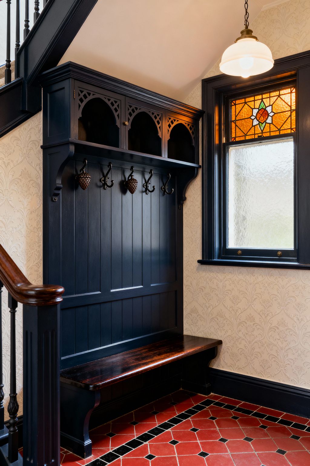 A Victorian-style mudroom corner features a deep navy built-in hall tree with arched cubbies, paneled back, and black metal acorn hooks above a dark-stained wood bench. Cream damask wallpaper lines the walls, a terracotta red tile floor with a black border grounds the space, and a window with amber and green stained glass sits beside a brass-and-opal glass pendant light.