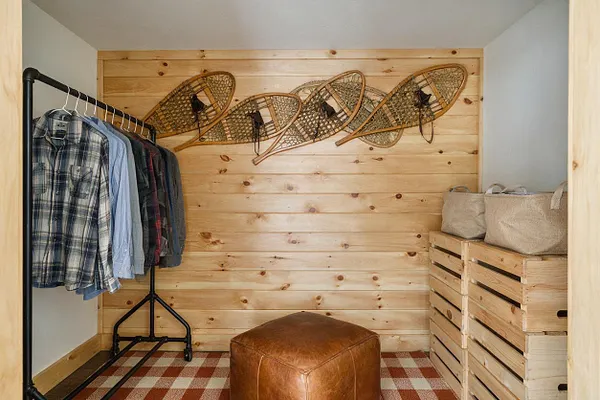 A narrow rustic closet features light knotty pine plank walls, a black metal clothing rack with hanging plaid and solid shirts, and stacked wooden crates on the right topped with woven storage baskets. Vintage snowshoes are mounted on the back wall above a brown leather pouf, with a red-and-cream checkered rug on the floor.