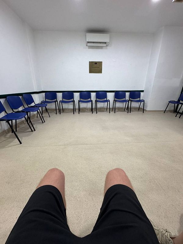 A waiting area for a driving theory test with empty blue chairs and a view of someone's legs.