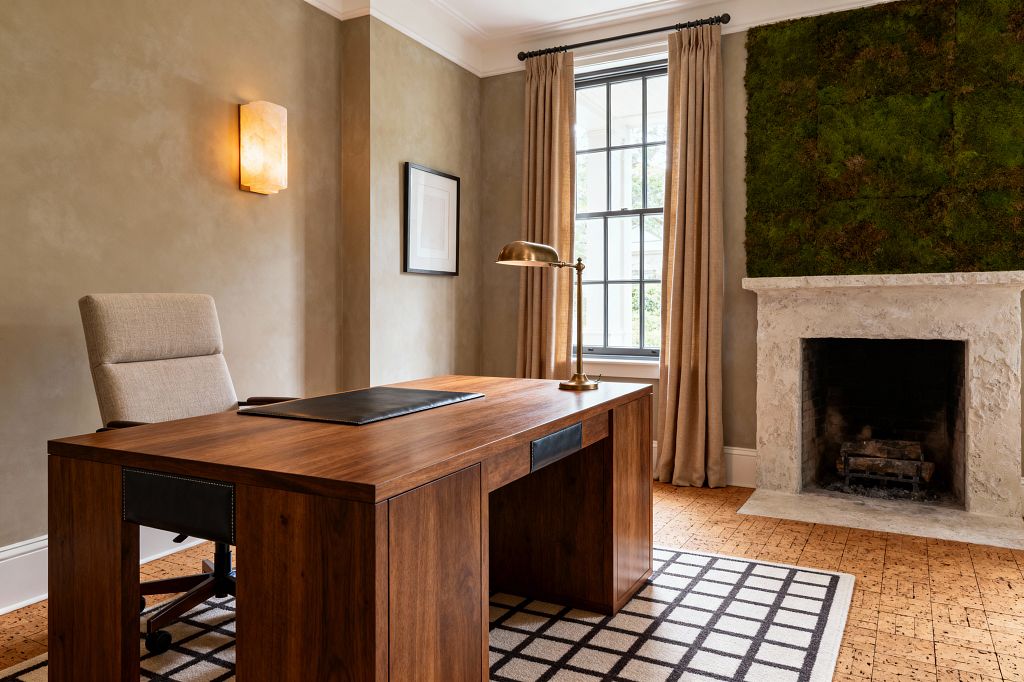 A transitional home office with warm taupe plaster walls features a rectangular walnut desk with a brass task lamp and a beige upholstered swivel chair. A large window with black muntins is dressed in tan linen curtains, while a textured stone fireplace is topped by green preserved moss panels. Cork flooring holds a cream rug with a black grid, and an alabaster sconce glows beside minimal framed art.