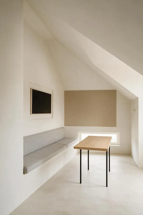 A minimalist game room with sloped white ceilings, a built-in light gray upholstered bench along the left wall, a small wood-top table with black metal legs in the center, a wall-mounted black screen, and a large beige acoustic panel on the back wall. The room is finished in soft neutral tones with daylight entering through a low window.