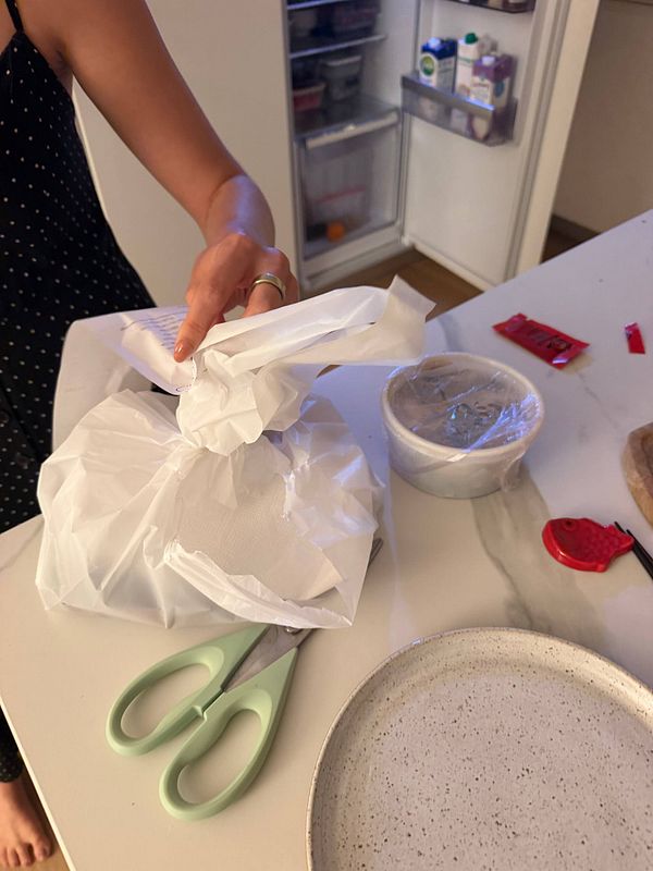 A person holding a wrapped food package on a kitchen counter with various items including scissors, a covered bowl, and a plate nearby.