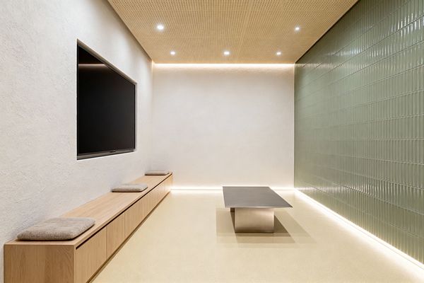Minimalist media room with a recessed flat-screen TV on a warm white textured plaster wall and a long, low built-in oak bench with handleless storage and three beige felt cushions. A glossy sage-green tiled wall lines the right side; a low brushed stainless-steel table sits on a seamless pale-beige floor. Perforated wood ceiling panels with recessed downlights and soft LED perimeter lighting create calm, even illumination.