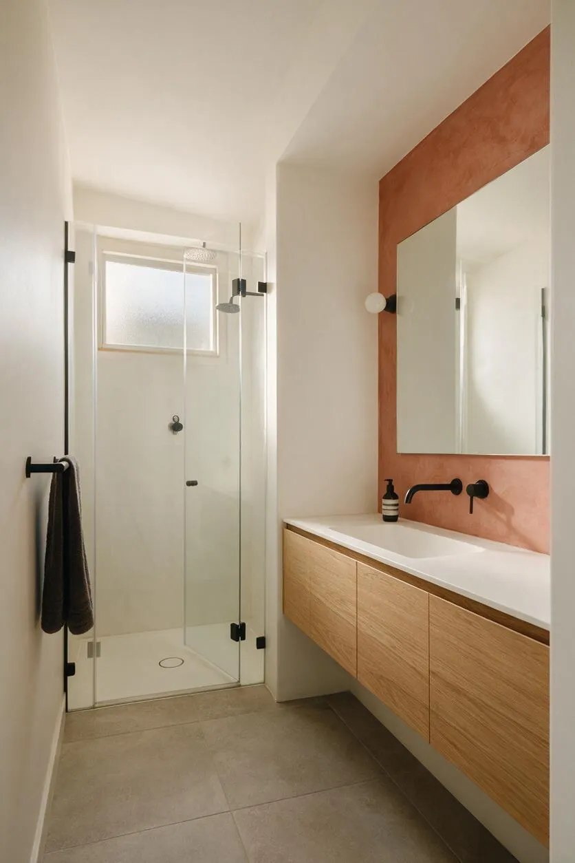A modern master bathroom with a frameless glass shower on the left and a floating light wood vanity on the right. The vanity has a white countertop, black faucet, and a large mirror set into a terracotta-toned accent surround, with neutral tile flooring and soft natural light from a small frosted window.