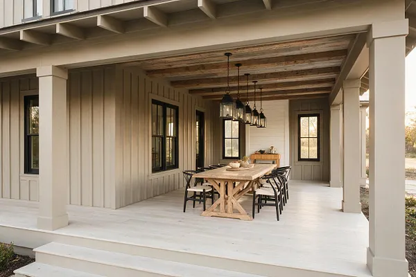 A spacious farmhouse front porch with a white painted floor, beige vertical board-and-batten siding, and exposed wood ceiling beams. A long rustic wood dining table sits in the center with black chairs, while black-framed windows and hanging lantern lights add contrast.