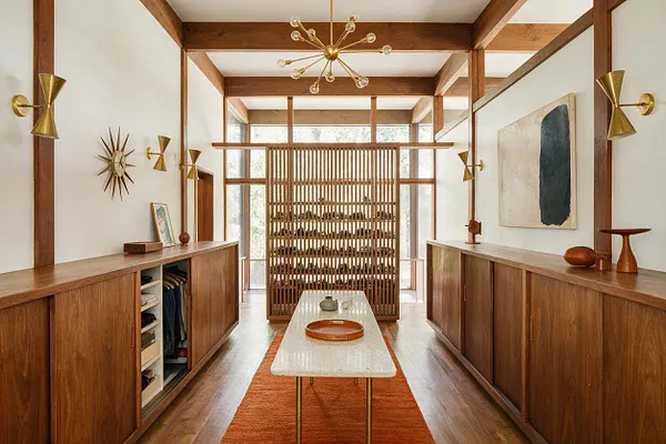 A long, symmetrical closet interior with walnut wood cabinetry on both sides, open shelving and hanging storage, and a central slatted divider at the back. Brass starburst wall sconces and a modern brass chandelier add Mid-Century Modern character, while a pale stone table and rust runner soften the warm wood tones.